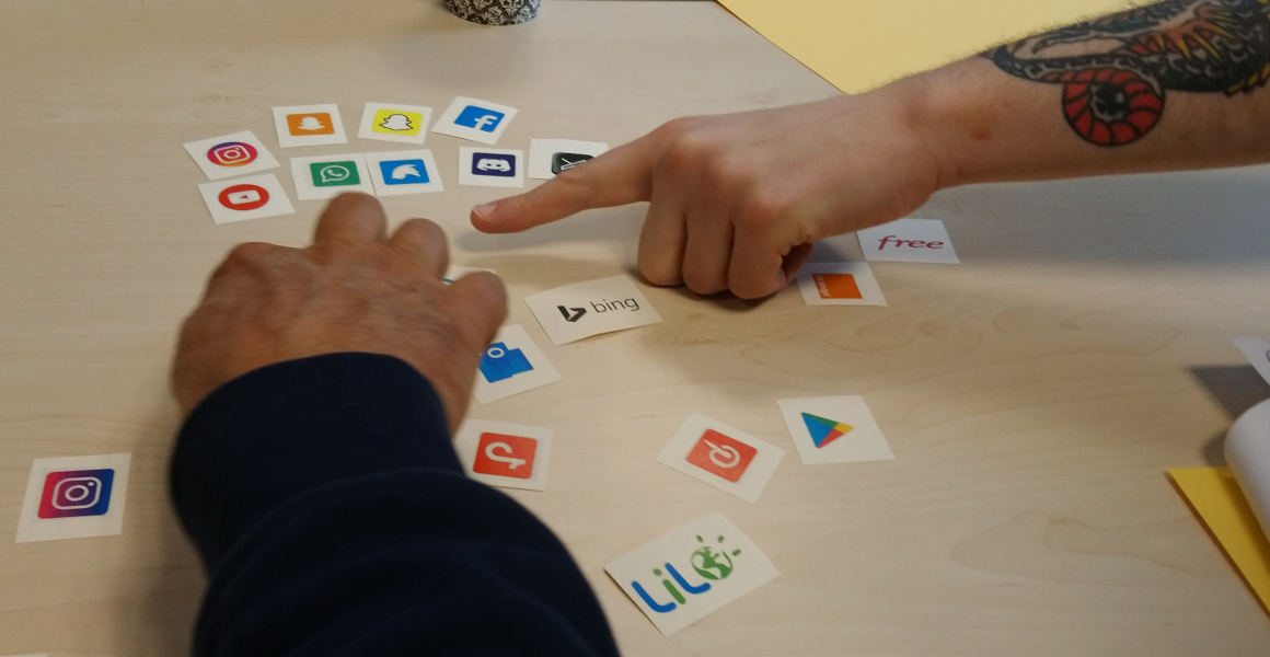 sur une table sont disposées plusieurs icônes de services numériques. Un bras tatoué pointe avec un index une de ces icône, pendant qu'un autre bras en saisit une autre. 